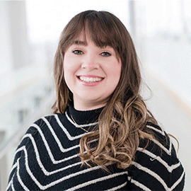 A woman wearing a stripped cardigan stands in front of a university corridor and smiles at the camera.